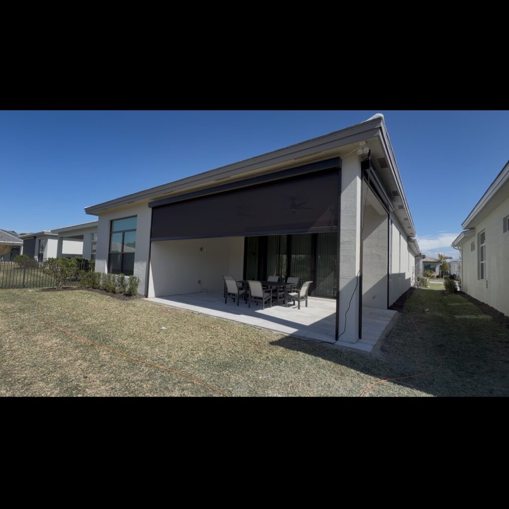A modern covered patio with a black retractable screen, outdoor dining table, and chairs, attached to a single-story house, overlooking a grassy backyard under a clear blue sky.