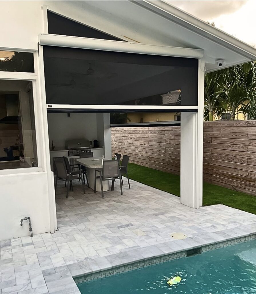 Outdoor patio area with a dining table and chairs, covered by a roof and enclosed with a retractable screen. The space is next to a swimming pool and bordered by a wooden privacy fence and some palm trees.
