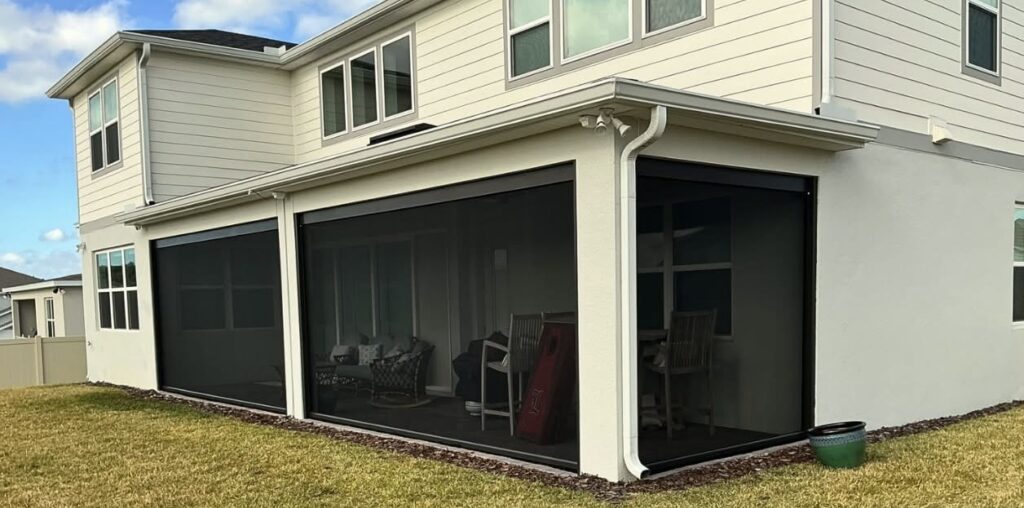 A modern two-story house with a screened-in patio area, outdoor chairs, and a small table. The house has white siding and multiple windows, with a lawn and a potted plant near the corner.
