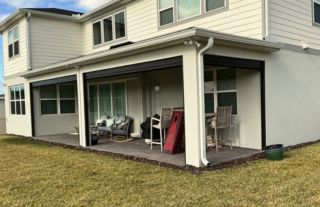 A covered backyard patio attached to a two-story house, featuring outdoor furniture, a cornhole game board, and a green planter, overlooking a grass lawn.