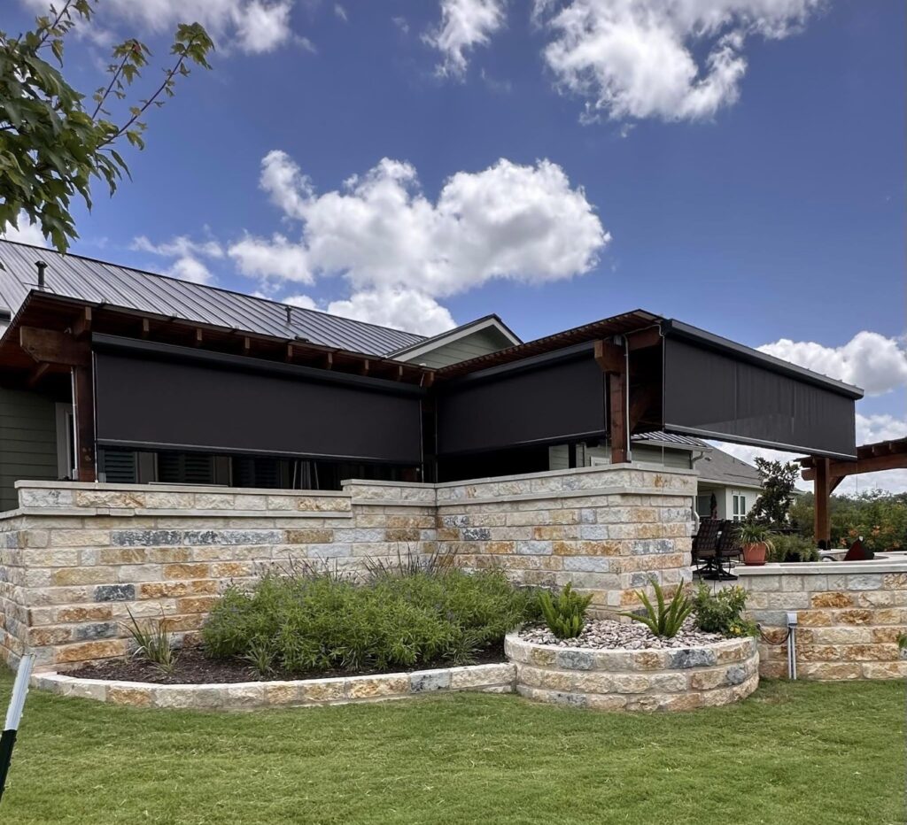 A modern house with stone walls and raised garden beds features retractable black outdoor shades on a covered patio, under a blue sky with scattered clouds.