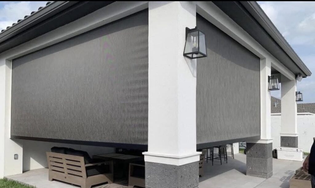 A covered outdoor patio area enclosed with large roll-down screens, featuring outdoor seating and a bar area, all under a modern dark roof with white pillars and wall-mounted lantern lights.