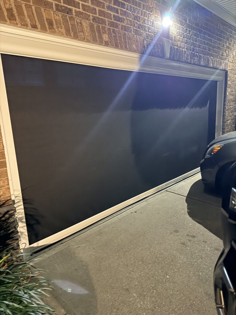 A closed garage door at night is shown, illuminated by an exterior light. Part of a parked black vehicle is visible on the right, and some plants can be seen in the lower left corner.