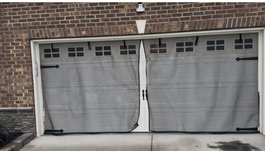 A closed double garage door with four small windows on each side is covered by a gray mesh screen, attached with black fasteners. The house is made of brick and has a stone accent on the left.