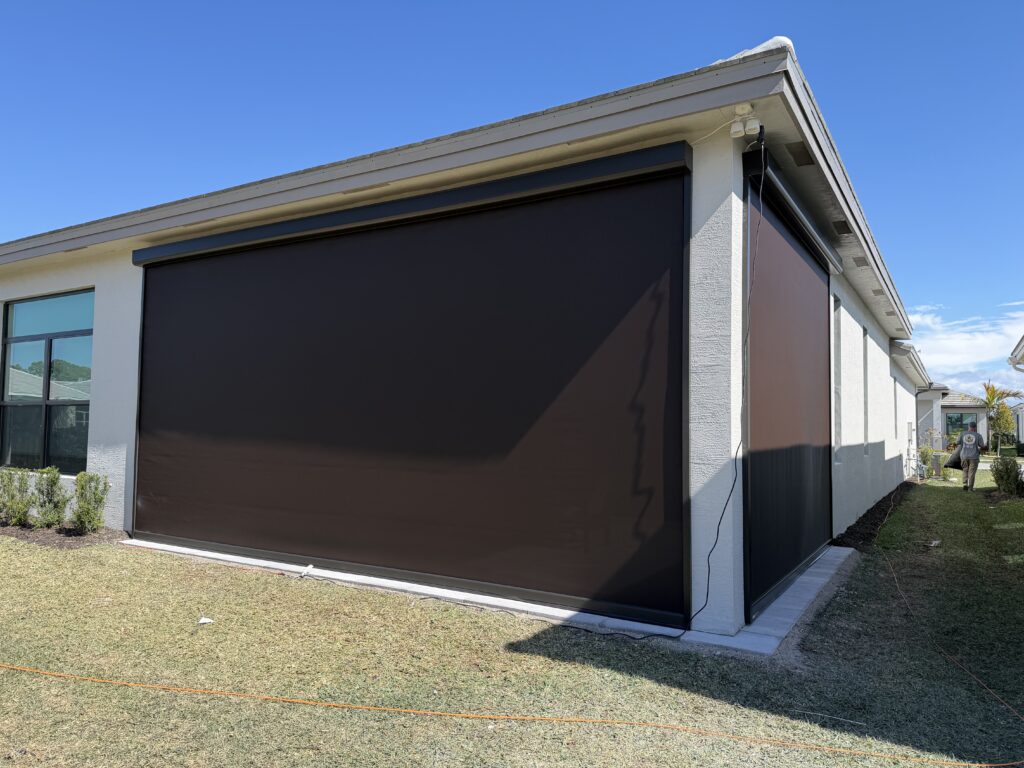 A house with large black motorized outdoor shades covering its patio openings on two sides, providing shade and privacy. The house sits on a grassy yard under a clear blue sky.