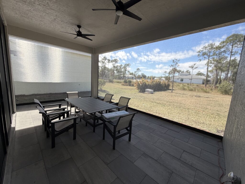 A screened-in patio with a rectangular table and eight chairs, ceiling fans above, and a view of a grassy yard with trees and shrubs under a blue sky.