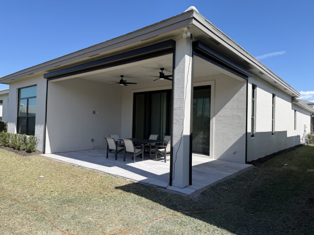 A modern house with a covered patio features two ceiling fans, a dining table with six chairs, and retractable screens. The patio opens to a grassy backyard under a clear blue sky.