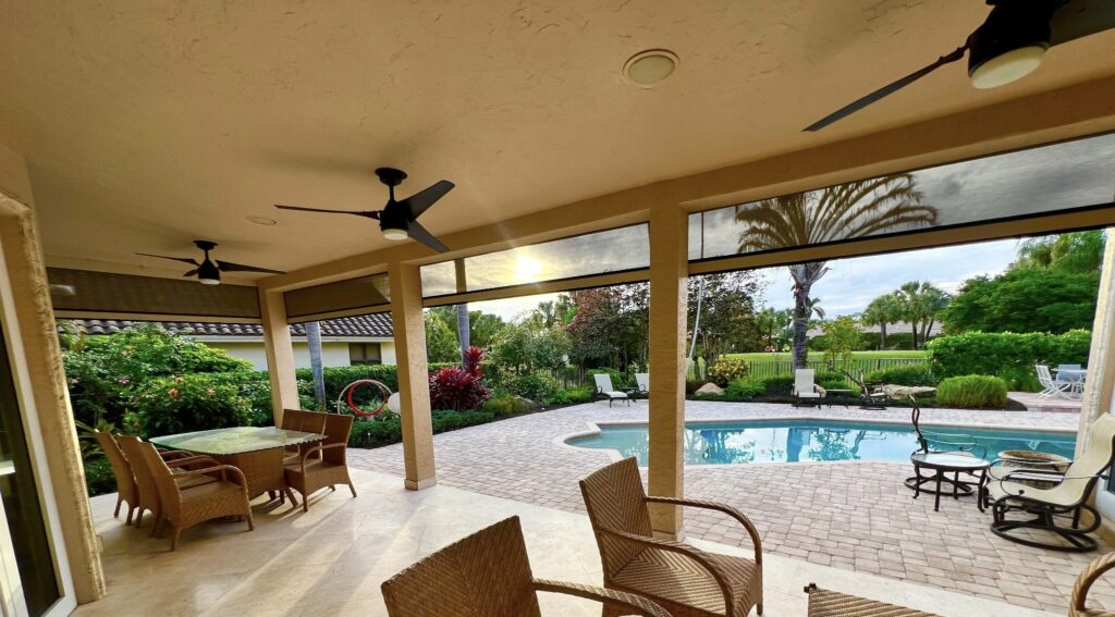 A covered patio with ceiling fans, wicker chairs, and a table overlooks a backyard with a pool, lounge chairs, lush greenery, and a golf course view at sunset.