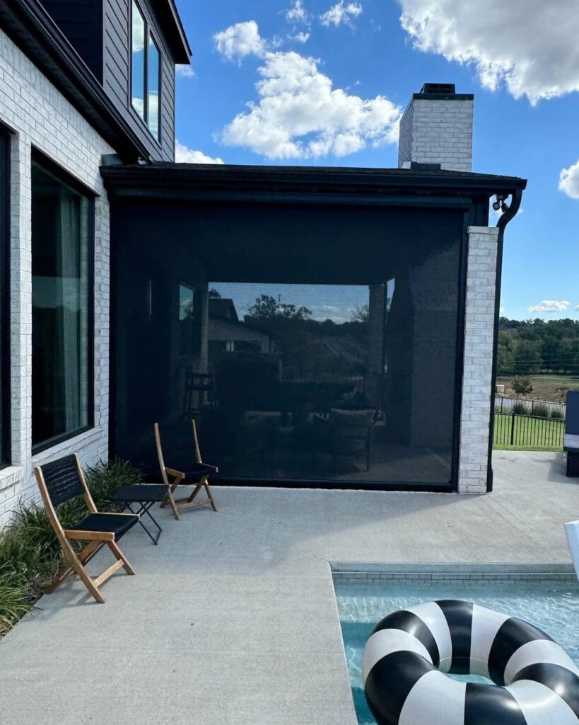 A modern house with a white brick exterior, a black mesh screen enclosing a patio area, two black chairs with wooden frames, and a pool with a black-and-white striped float nearby. Trees and clouds are in the background.