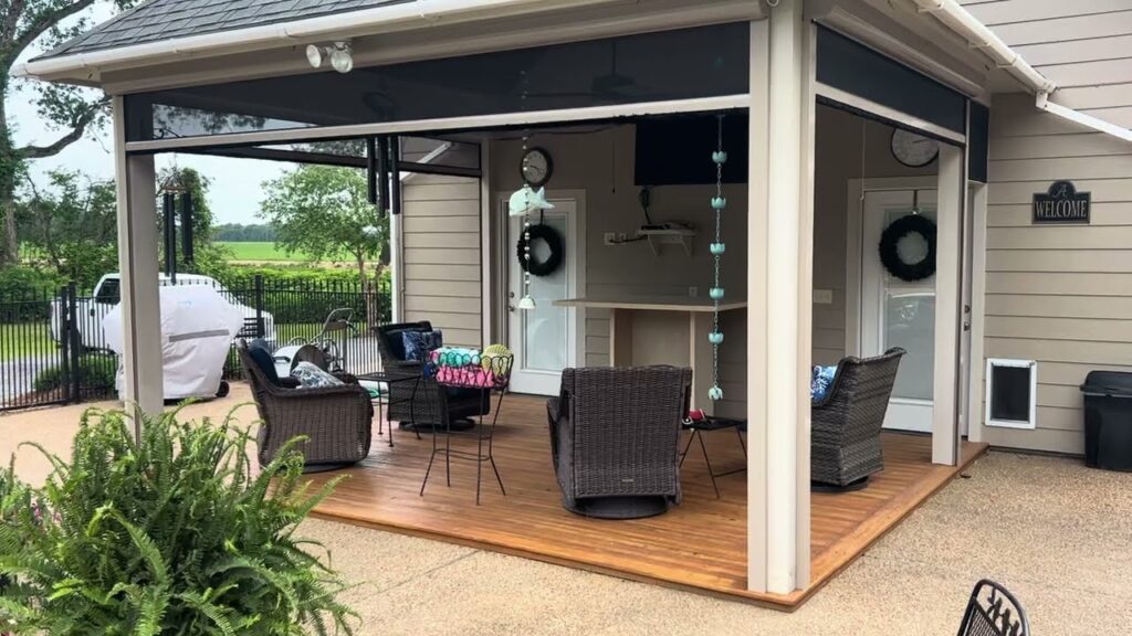 A covered outdoor patio with wicker chairs, a small table, hanging lights, two doors with wreaths, a TV, and a bar area. The space overlooks a yard with green trees and a white grill in the background.