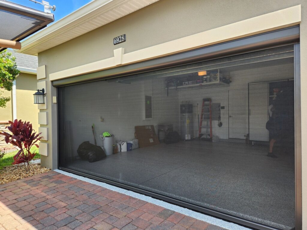 A closed, transparent garage door reveals a clean, organized garage with bins, bags, shelves, and a folded ladder inside. The exterior features beige walls, pavers, and some landscaping with red plants.