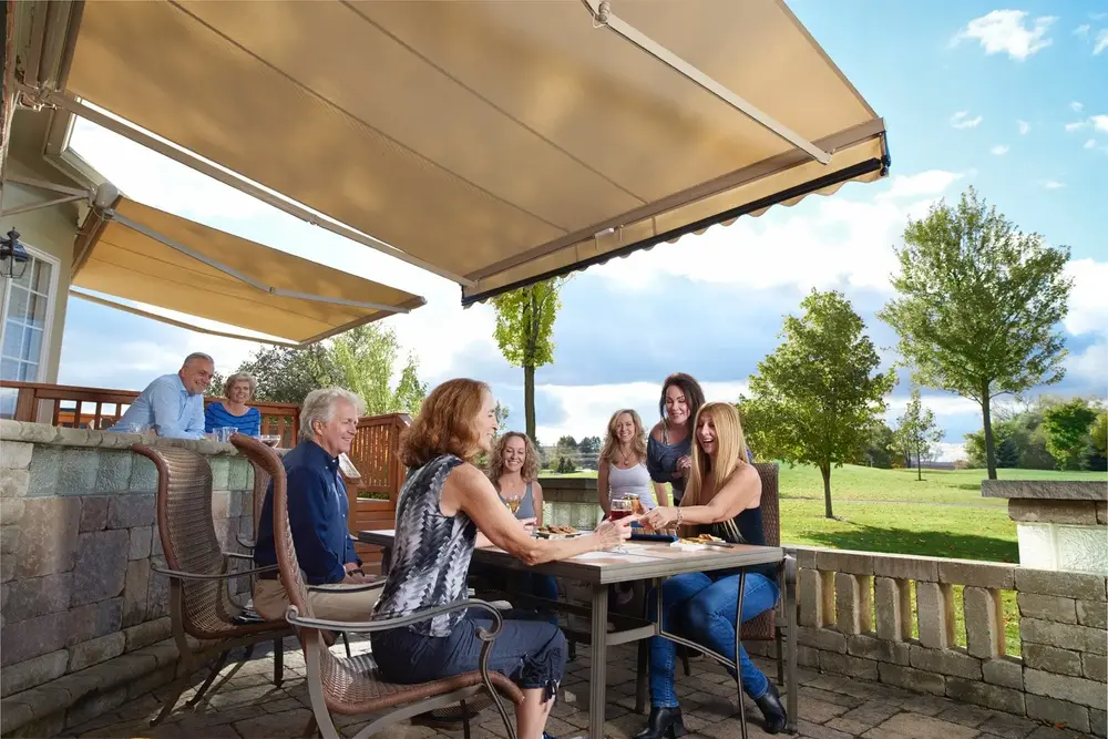 A group of people gather around a table on a sunny patio with large retractable Sunpro awnings overhead. Some are sitting, some standing, and they appear to be enjoying drinks and conversation with a view of green trees and an open field.