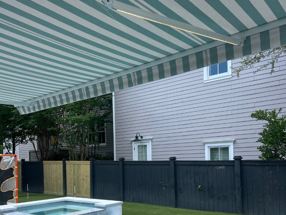 A striped blue and white Sunpro retractable awning extends over a backyard with a small pool, black fence, and a light purple house with white-framed windows in the background.