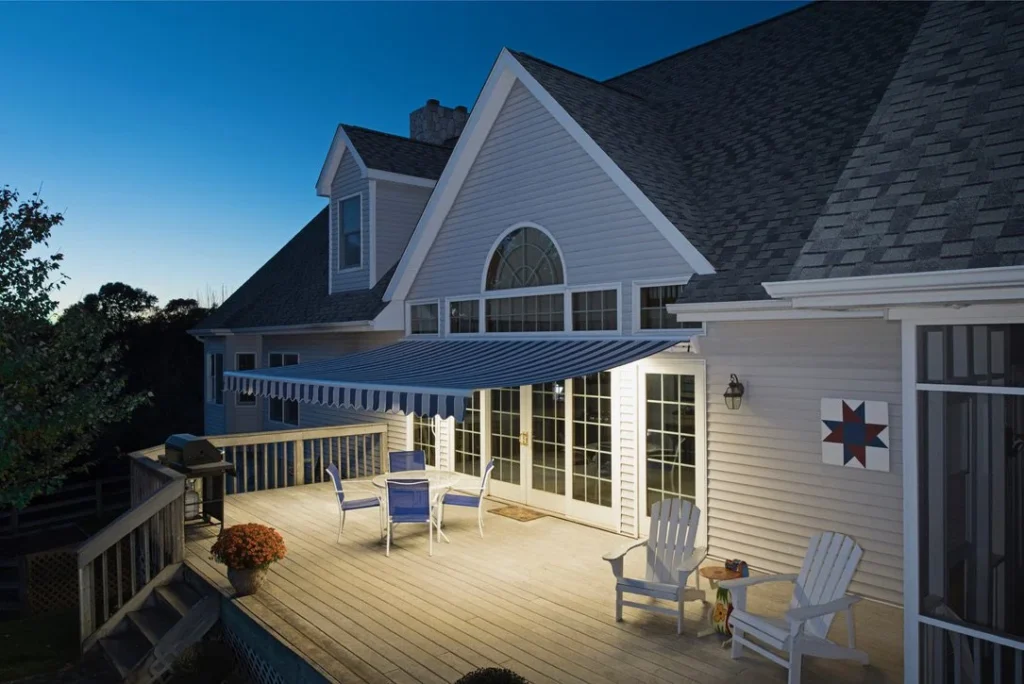 A spacious wooden deck attached to a house at dusk, featuring white Adirondack chairs, a table with blue chairs, potted plants, a grill, and a blue-striped retractable awning from SunPro and Simpli Shaded.
