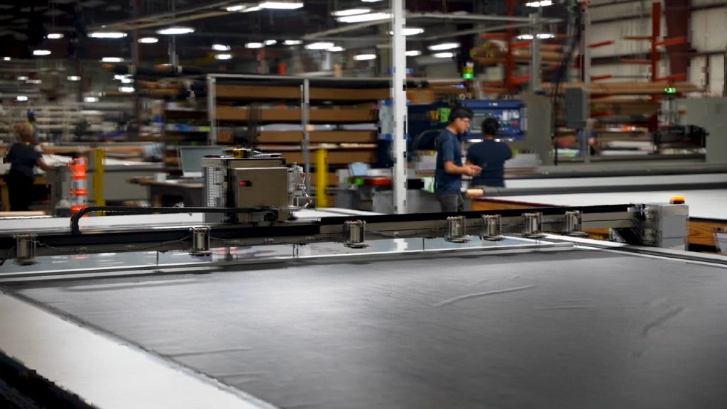 A large SunPro automated fabric cutting machine operates in a spacious industrial factory, with workers in the background attending to various tasks and shelves filled with materials.