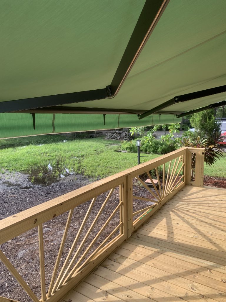 A wooden deck with a decorative railing sits under a large green retractable awning, overlooking a grassy yard with some puddles and tropical plants in the background.