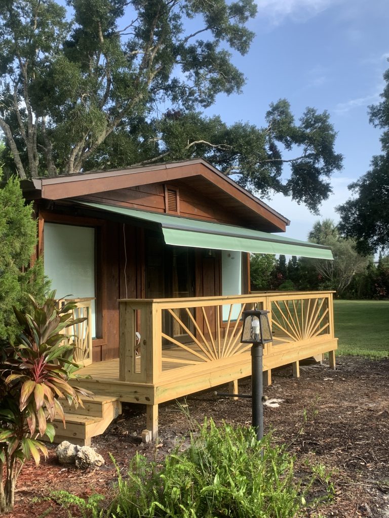 A small wooden cabin with a green awning and sunburst-patterned railing sits in a landscaped yard, surrounded by greenery and trees under a blue sky.