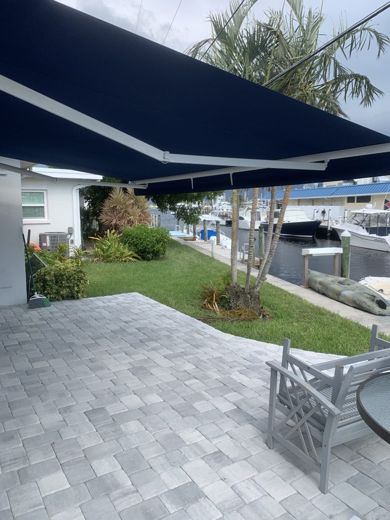 Patio with gray pavers, gray outdoor furniture, and a round glass table under a navy awning, overlooking a canal with boats and palm trees in the background.