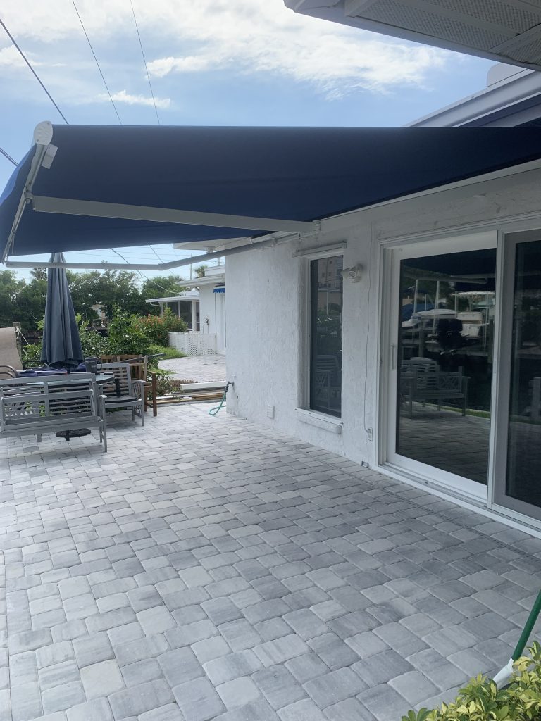 A patio with gray pavers features a blue retractable awning attached to a white house with sliding glass doors. There is a patio table and umbrella in the background, and greenery along the side.