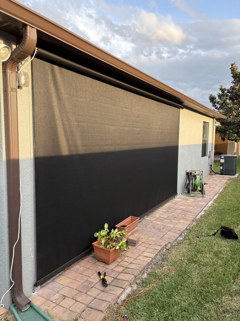 A black mesh shade covers a patio area attached to a house. There are two rectangular flower pots, a drill, a green garden hose, and an orange extension cord on the brick walkway. Grass borders the walkway.