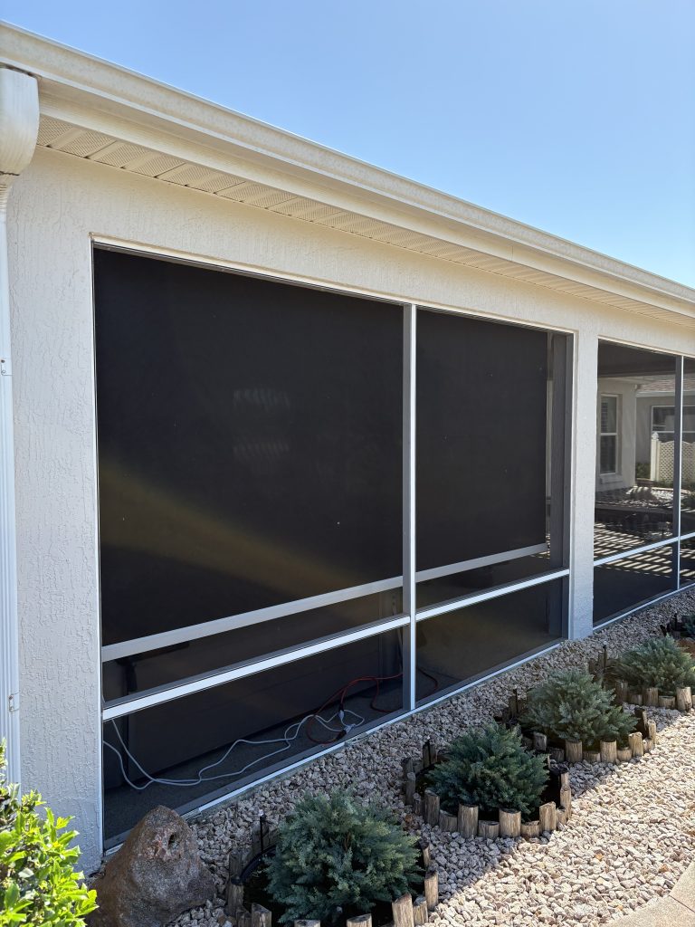 A screened-in porch with black mesh screens on a white house, surrounded by a rocky garden bed with small green shrubs bordered by short wooden edging, under a clear blue sky.