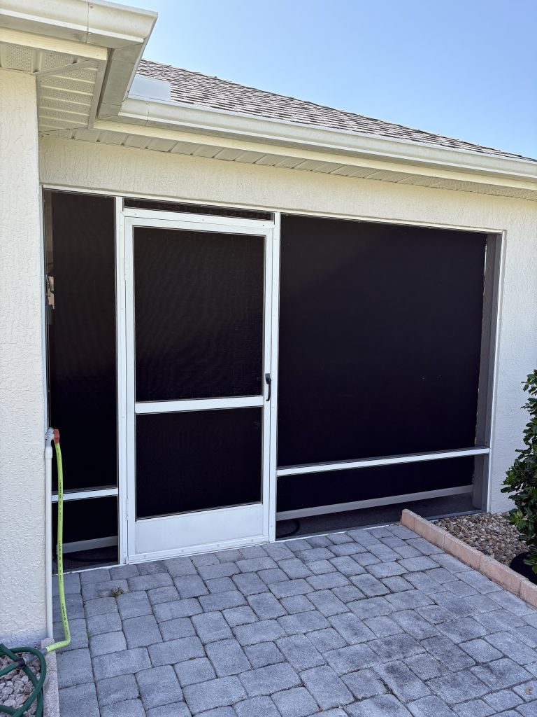 A screened patio door and two large screened panels attached to a light-colored house, with a paved stone patio and a green garden hose on the left side. Shrubs and a small raised garden bed are visible on the right.