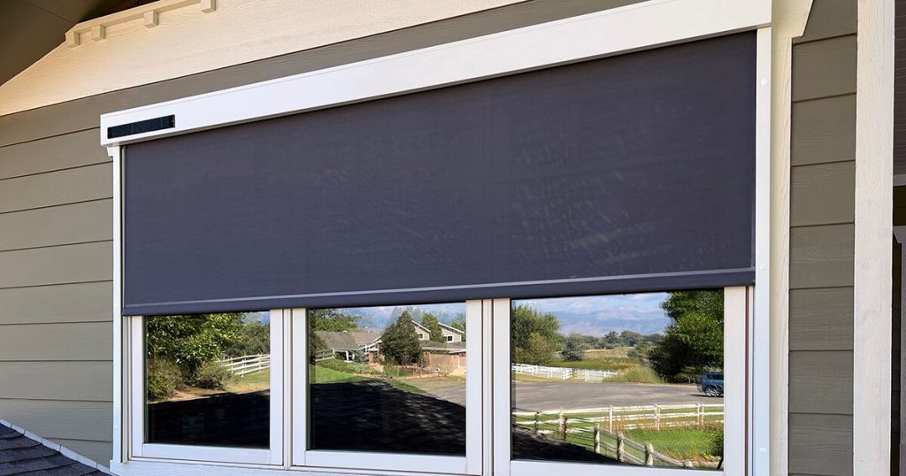 A large window with a black outdoor roller shade is mounted on the exterior wall of a house, partially covering three glass panes and reflecting a view of a rural landscape with trees and a road.