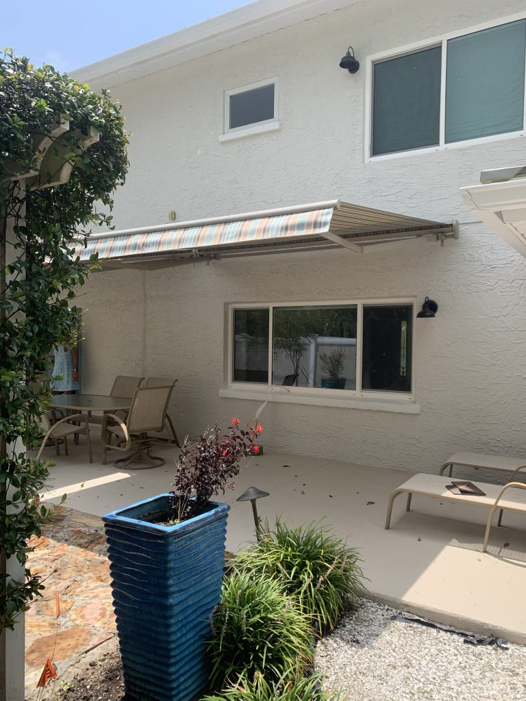 A backyard patio with a striped retractable awning, outdoor dining table, chairs, lounge chair, and a tall blue planter with plants. The patio is attached to a light-colored stucco house with two windows.