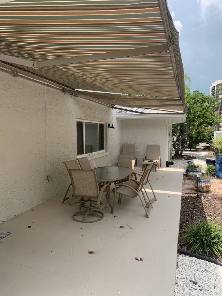 A covered patio with a striped retractable awning, beige outdoor table and chairs, and a window on a white stucco wall, overlooking a landscaped garden with gravel and plants.