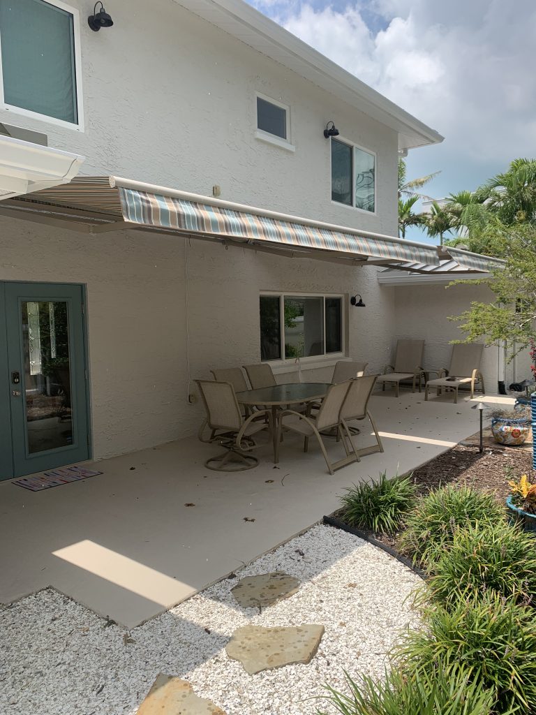A covered patio with a long outdoor dining table and chairs is attached to a two-story house. The area is shaded by a striped retractable awning, with plants and stepping stones in the foreground.