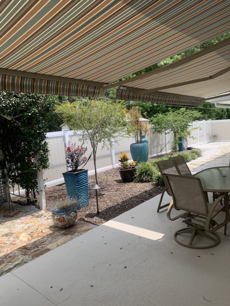 A backyard patio with a striped retractable awning, outdoor dining table and chairs, potted plants, small trees, and a white fence enclosing a landscaped garden area.