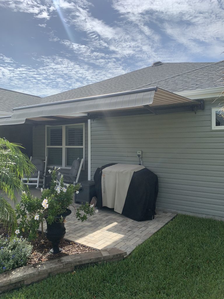A covered barbecue grill sits on a paved patio against a gray-sided house, with a retractable awning overhead. A potted plant and some shrubs are nearby, and the sky is partly cloudy.