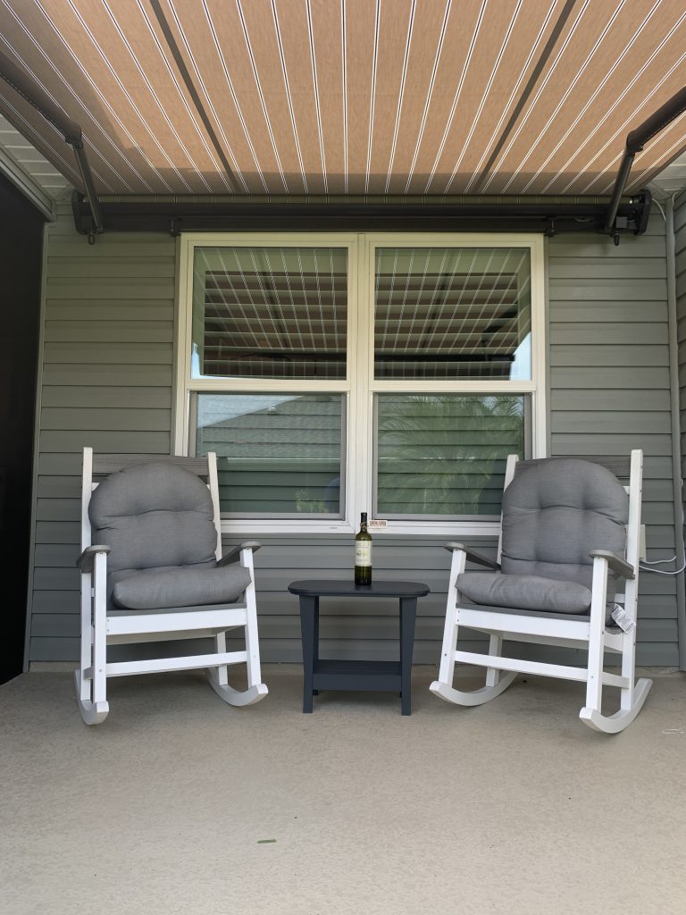 Two cushioned rocking chairs face each other on a porch beneath a striped awning, with a small table holding a bottle between them, in front of double windows on a gray-paneled wall.