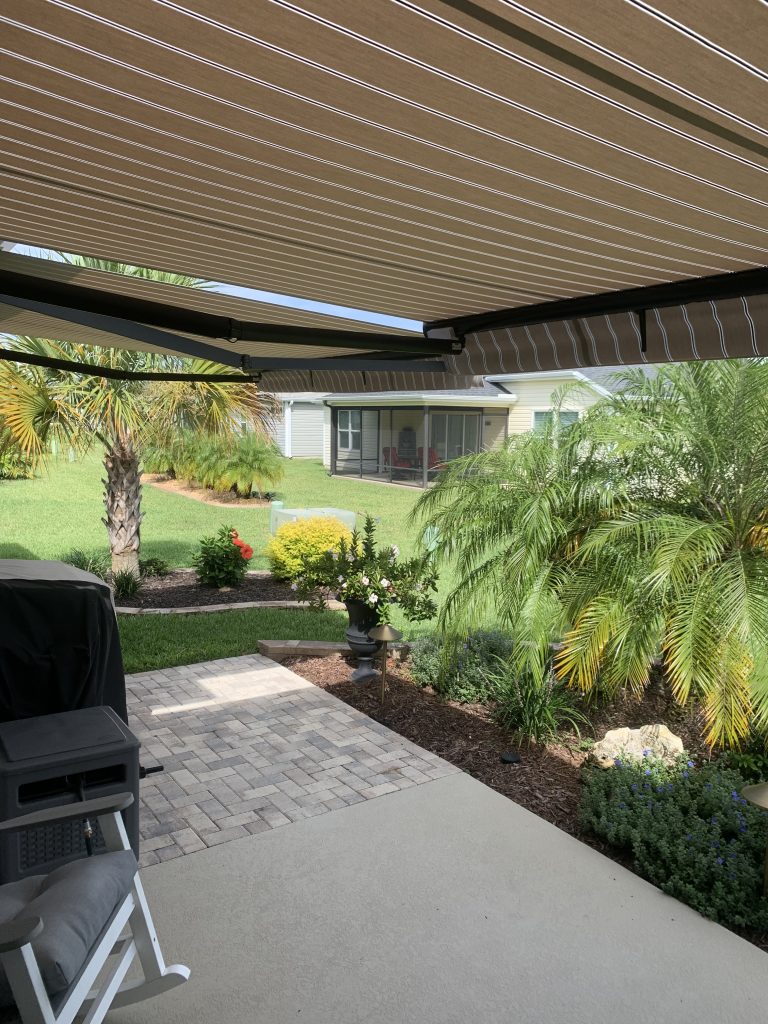 A shaded patio with striped awning overlooks a green backyard with palm trees, a flower garden, and a small building with a screened porch in the background. A chair and grill are visible in the foreground.