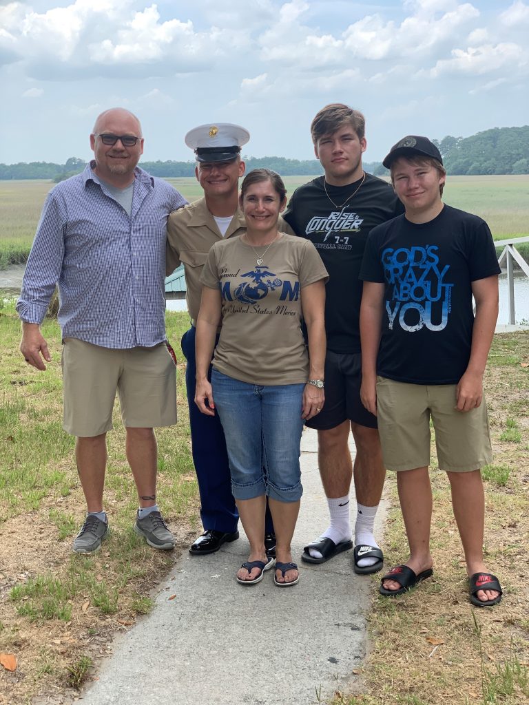 A smiling Boval family of five stands outdoors on a path near grassy water. One person wears a Marine uniform, while the others wear casual summer clothes. The background shows trees, open sky, and a dock by the water.