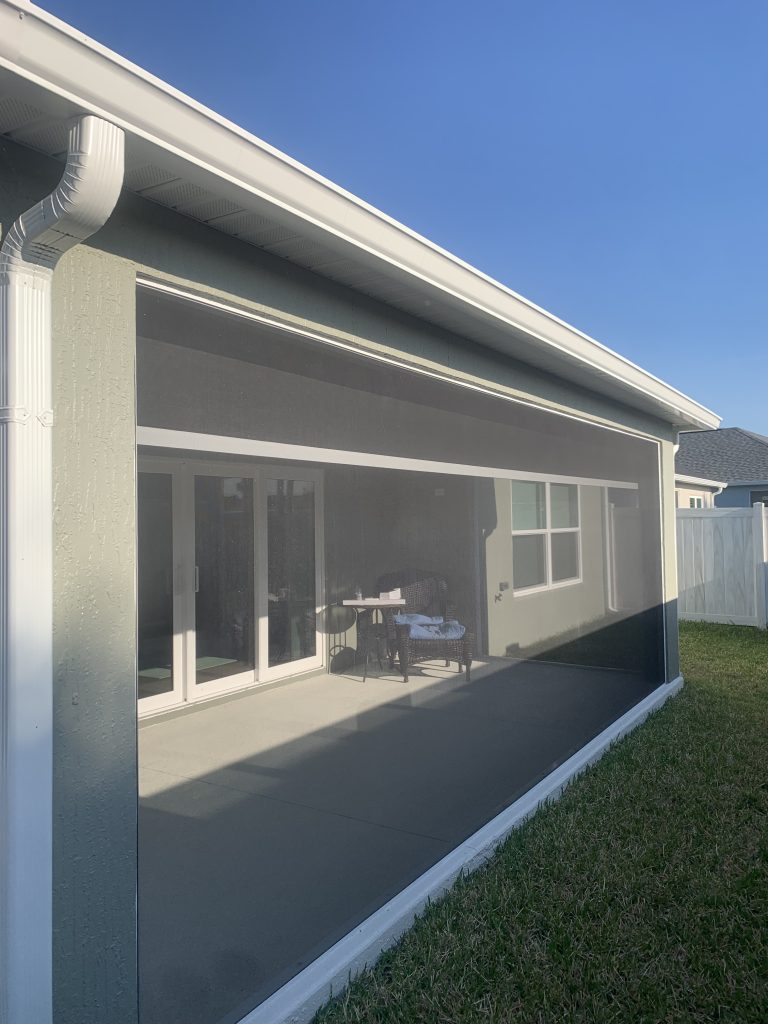 A screened-in porch attached to a house with sliding glass doors, seen from outside. The porch area is shaded, and there is green grass in the foreground under a clear blue sky.