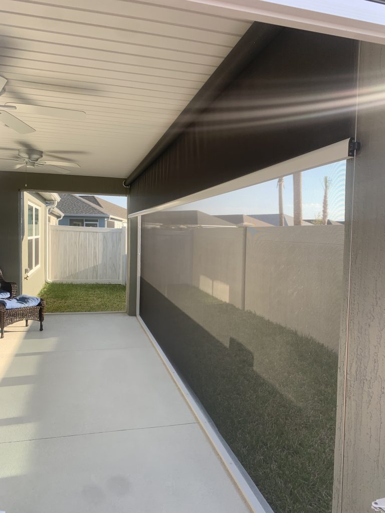A covered patio with a ceiling fan, enclosed by a transparent roll-down screen, overlooks a fenced backyard with grass and a neighboring house visible in the background.