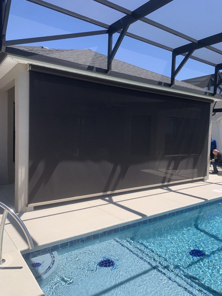 A screened patio with a lowered dark mesh shade overlooks a swimming pool in a sunny backyard, with clear blue sky and geometric shadows on the ground.