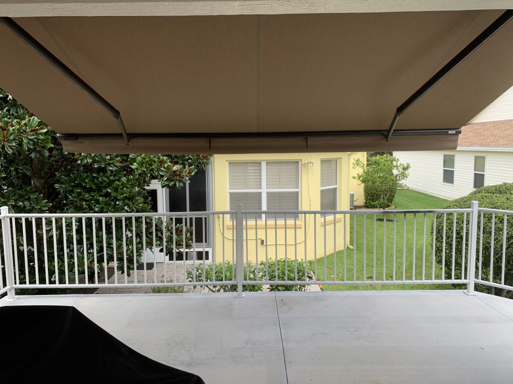 A covered patio with a brown retractable awning overlooks a white railing and green lawn. In the background, there is a yellow house with large windows and another white house nearby.