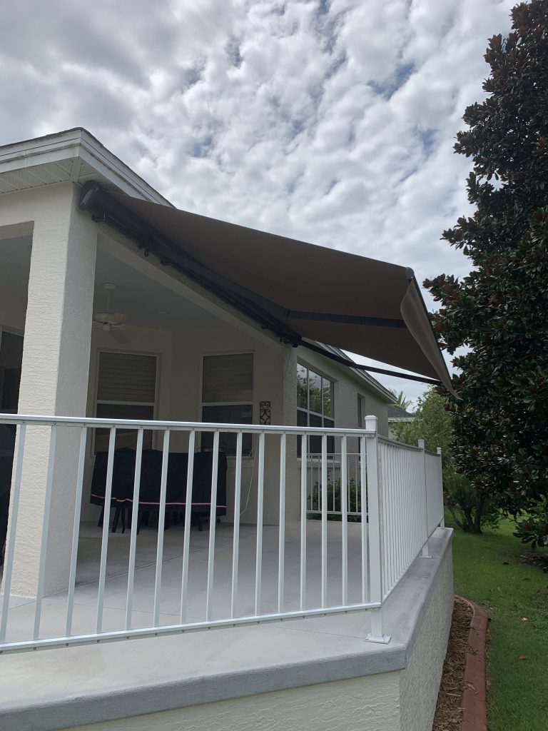 A white house porch with a white railing and a brown retractable awning extended overhead. The porch is empty, and the sky is mostly cloudy. Trees and greenery are visible in the background.