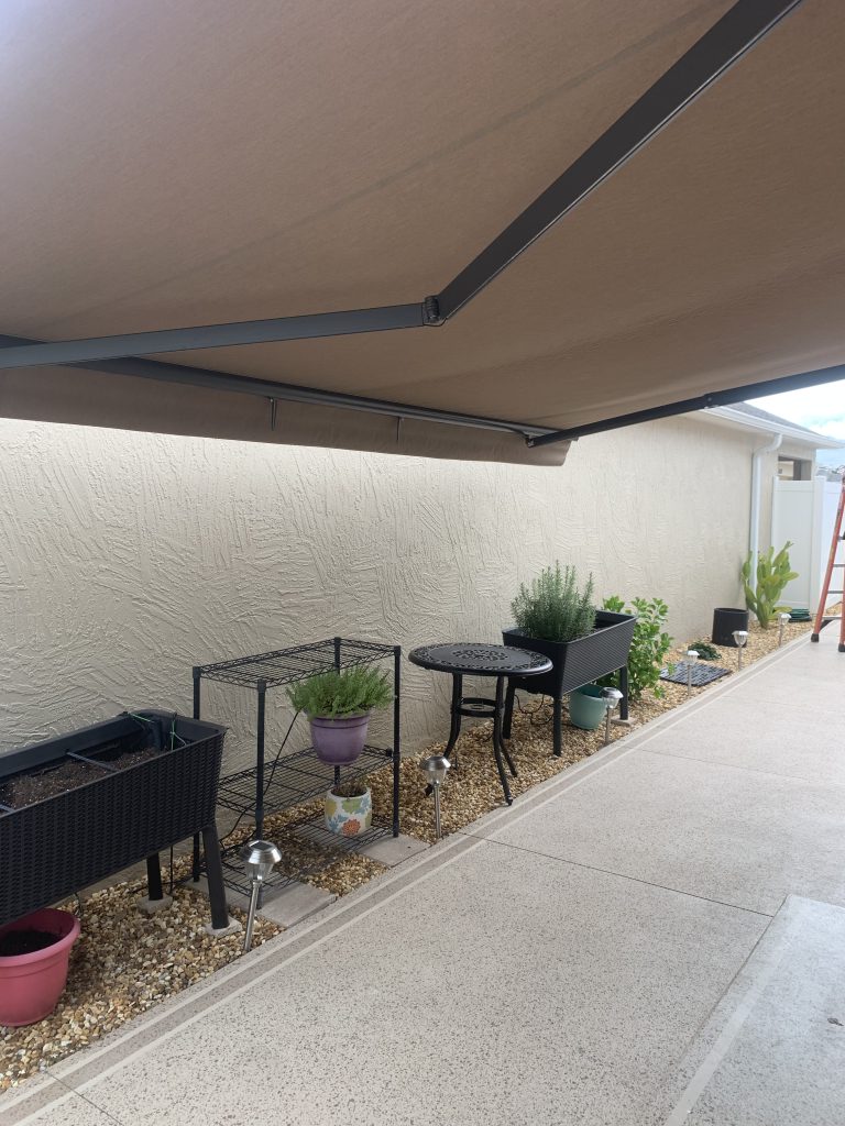 A covered patio area with a retractable awning, small black metal table and chairs, potted plants, planter boxes, gravel landscaping, and a white textured wall. A ladder and more plants are visible in the background.