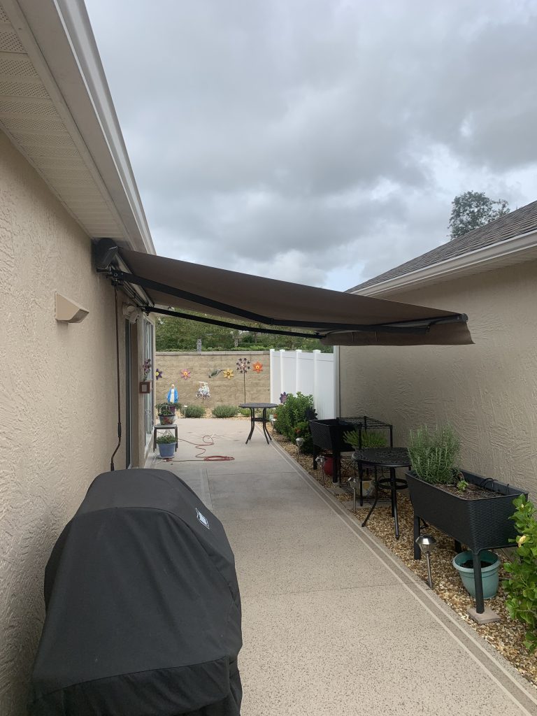 A retractable awning extends over a narrow patio between two beige buildings, with a covered grill, plants, and patio furniture beneath. The sky is cloudy, and a fenced yard with decorations is visible in the background.