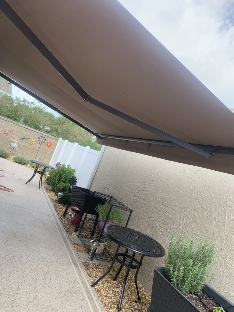 A covered patio area with a brown retractable awning, black metal tables and chairs, potted plants, and a textured beige wall with colorful flower decorations. White fence and greenery are in the background.