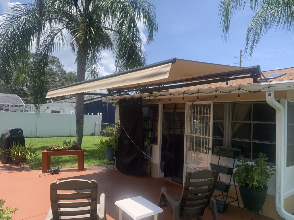 A beige retractable awning extends over a patio attached to a house. Patio chairs, a table, and potted plants are arranged beneath and around the awning. Palm trees and a grassy yard are visible in the background.