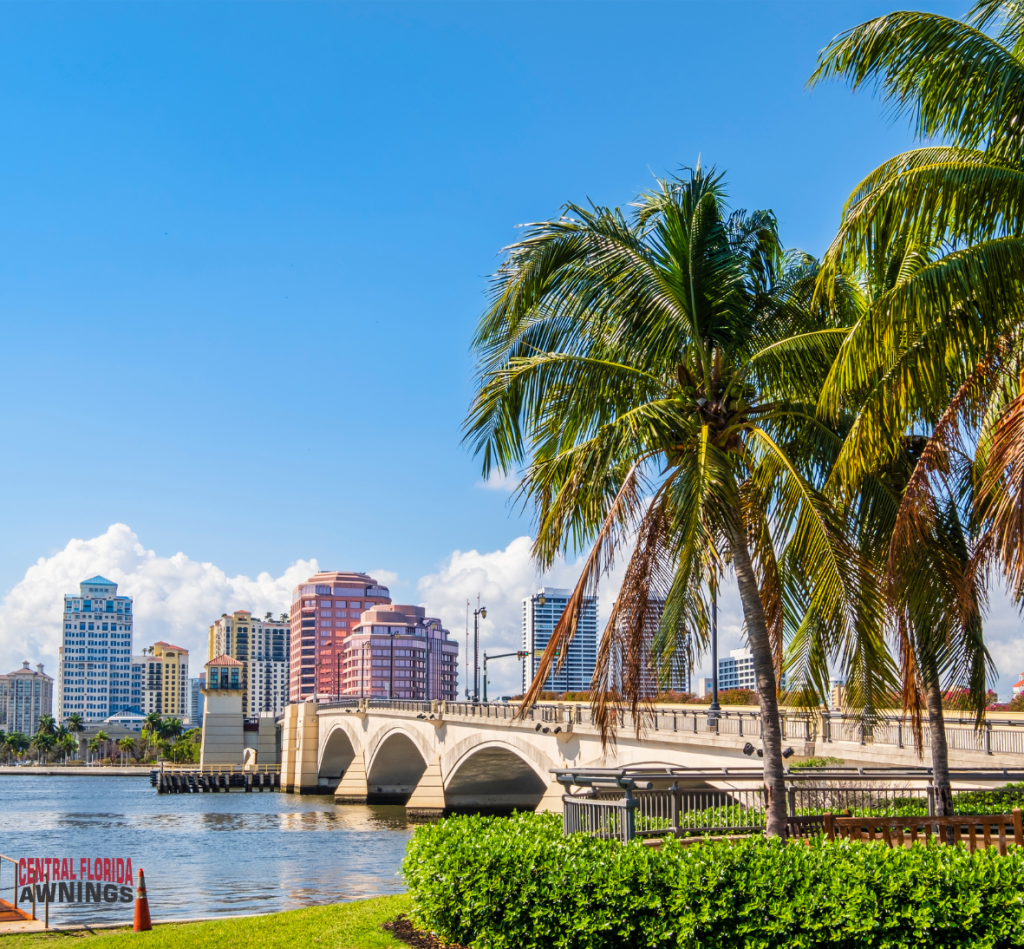 A scenic view of Spring Hill FL skyline with colorful high-rise buildings, a white arched bridge over the water, palm trees, green grass, and a blue sky with a few clouds. A Central Florida Awnings sign highlights retractable awnings Lakewood Ranch FL.