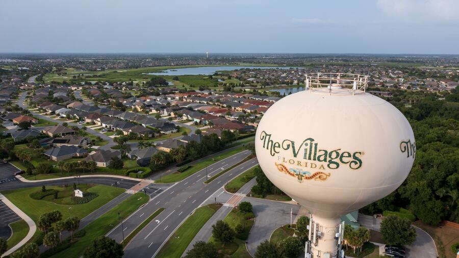 A large water tower labeled The Villages Florida stands above a sprawling residential neighborhood with rows of houses, green lawns, trees, and a pond in the background under a clear sky.