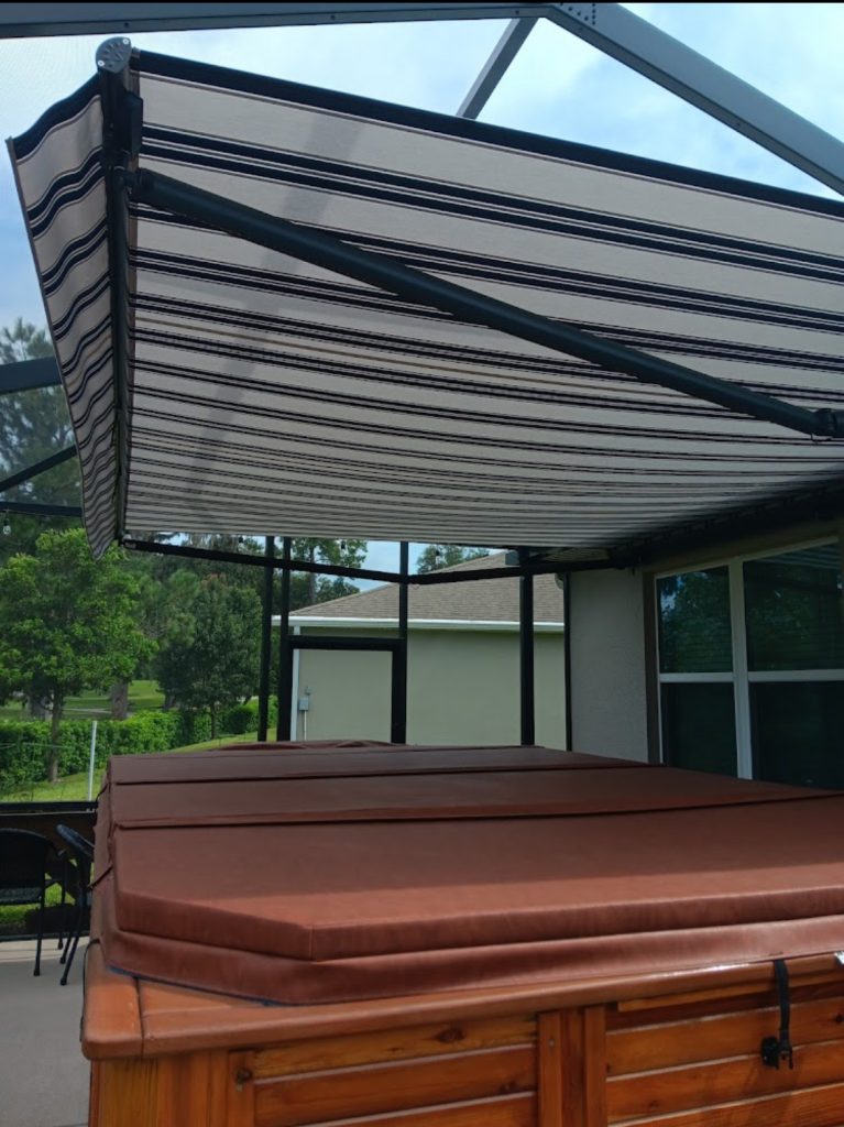 A brown hot tub with a closed cover sits on a patio beneath a large, extended striped canvas awning attached to the house, providing shade. Trees and a screened enclosure are visible in the background.