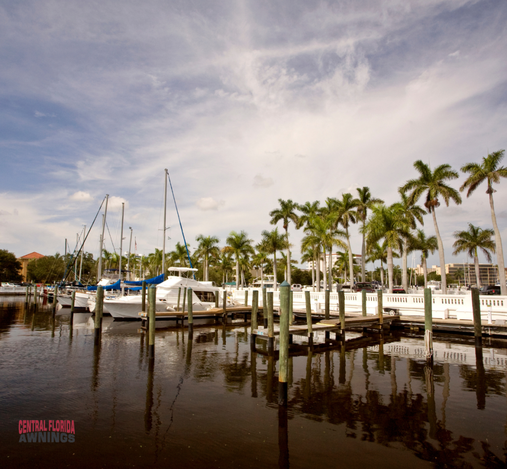 A marina near The Villages, Florida features docked sailboats and yachts lined with tall palm trees under a partly cloudy sky. Central Florida Awnings is visible in the lower left corner, reflected in the calm, dark water.