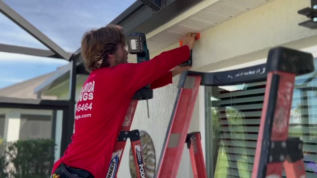 A person in a red shirt stands on a ladder, installing a small device under the eaves of a house near a window. Another ladder is positioned nearby. The scene appears to be outdoors on a sunny day.