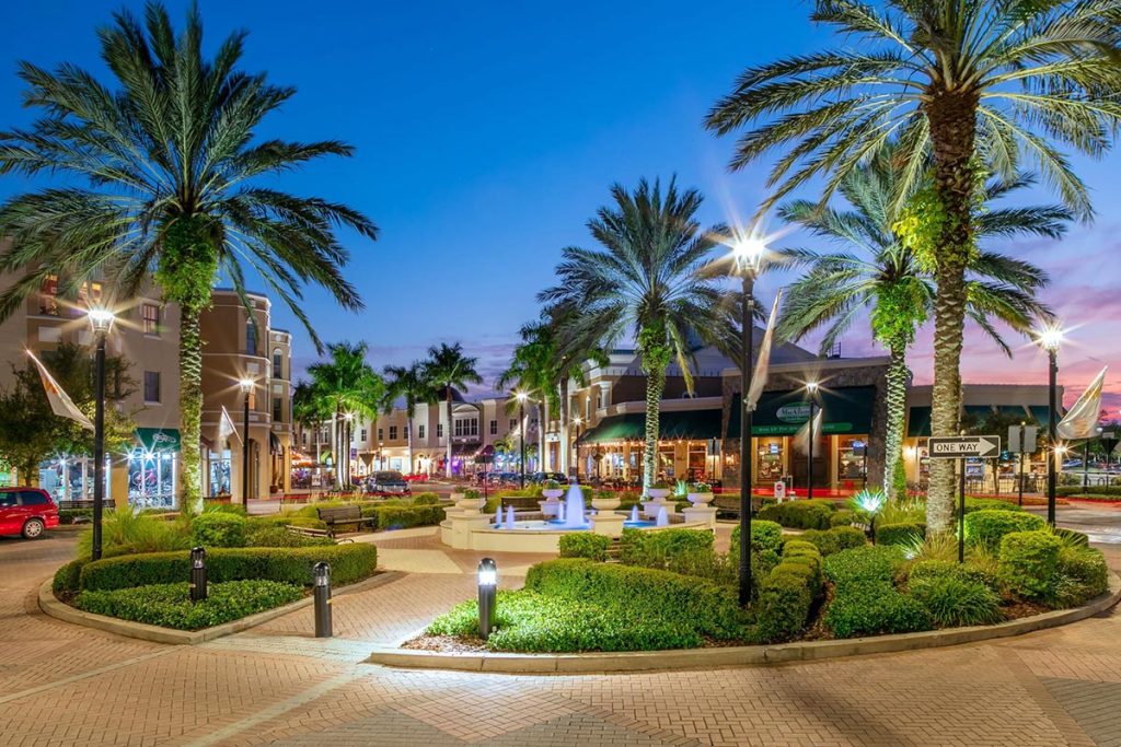 A vibrant Lakewood Ranch outdoor plaza at dusk in The Villages, Florida, with palm trees, manicured gardens, a central fountain, street lamps, awnings over surrounding shops and restaurants, creating a lively and inviting atmosphere.